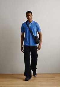 Blue polo shirt, black wide-leg pants, and black shoes. Crossbody bag with hardware detail. Model walking against a neutral backdrop.