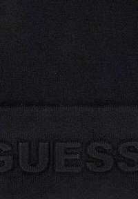 Tissu en maille noir avec une finition texturée, présentant un logo en relief proéminent qui indique "GUESS" en bas.