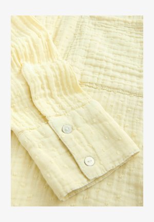 Hellgelber Hemdenstoff mit einem strukturierten Muster. Ausgestattet mit einem Ärmelabschluss und zwei weißen Knöpfen. Weiches, gewebtes Material mit erhabenen Punkten.