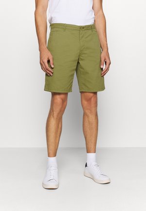 Olivgrüne Shorts aus Baumwolle, mit flachem Bund, Gürtelschlaufen und seitlichen Taschen. Kombiniert mit weißen Turnschuhen und Socken.