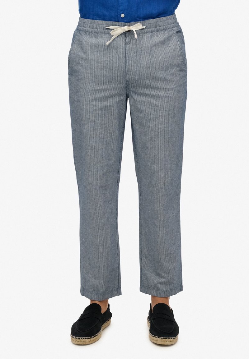 Lichte, blauw gestreepte broeken met een elastische tailleband, trekkoord en voorzakken. Gepaarde met zwarte instapschoenen.