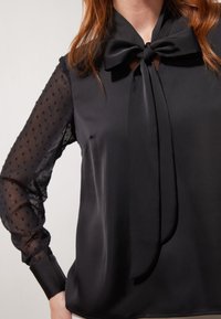 Black blouse with a satin body and sheer, dotted sleeves. Features a bow tie at the collar and buttoned cuffs. Smooth texture overall.