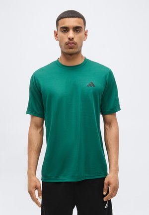 Mand iført en grøn kortærmet sportstrøje med et sort Adidas-logo, stående mod en ensartet lys baggrund.
