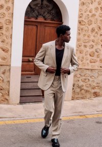 Homme en costume beige et chemise noire marchant dans la rue, ajustant sa veste, devant une porte en bois et un mur en pierre.