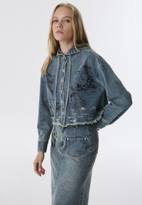 Gekürzte blaue Jeansjacke mit Kapuze, ausgestattet mit einem naturbezogenen Druck, fransigem Saum und Knopfverschluss; kombiniert mit einem passenden Jeansrock.