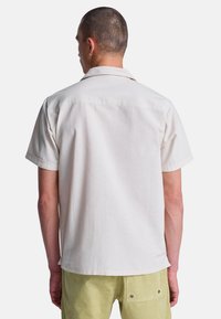 Hombre de espaldas con camisa crema de manga corta y pantalones verde claro contra un fondo blanco liso.