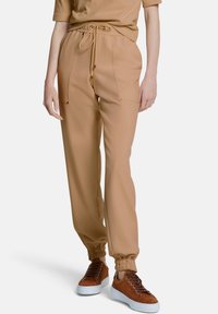 Beige joggings met elastische boorden, twee voorzakken en een trekkoord in de taille. Gecombineerd met bruine sneakers. Glad stofmateriaal.
