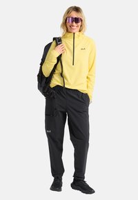 Maglia gialla a maniche lunghe con collo con zip, abbinata a pantaloni neri con tasche. Gli accessori includono occhiali da sole scuri e uno zaino nero.