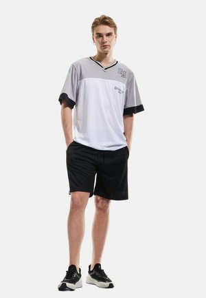 Sportsoutfit bestående af en hvid og grå kortærmet trøje med sorte detaljer, kombineret med sorte shorts og sportssko.