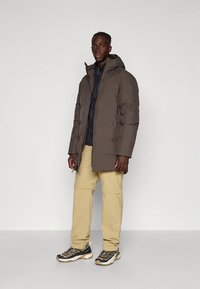 Smeđa puff jakna s kapuljačom, zatvaranjem na patentni zatvarač i bočnim džepovima, slojevito preko tamne majice i uparena s kremasto bijelim hlačama i sportskim cipelama.