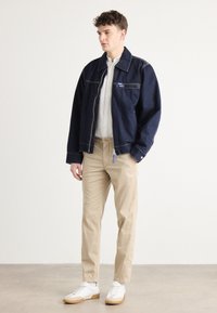 Tumši zili džinsi žakete ar lielām kabatām, gaišā krāsā pogājama krekla, bēšu bikses un baltas kedas ar gumijas zoli.