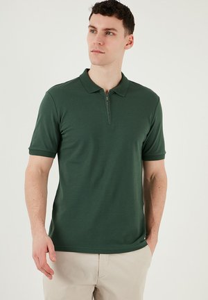 Dunkelgrünes Polohemd mit Reißverschlusskragen, kurzen Ärmeln und glatter Textur. Getragen mit beigen Hosen, die einen lässigen Schnitt zeigen.