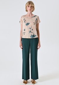 Top beige chiaro con stampa floreale, abbinato a pantaloni larghi verde scuro. L'outfit è completato da scarpe decorate con fiocchi dorati.
