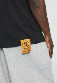 Melna T-kreklu ar oranžu birku, kurā ir "ff" logo, karājoties no sānu šuves, apvienota ar pelēkiem sporta biksēm.