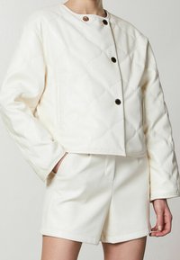 Giacca bianca trapuntata con scollatura rotonda, chiusura con bottoni e tasche laterali. Abbinata a pantaloni corti bianchi coordinati, entrambi realizzati in materiale morbido.