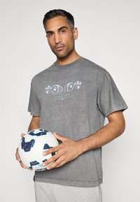 Γκρι βαμβακερό μπλουζάκι με αντίθετο γραφικό τύπωμα στο μπροστινό μέρος, σε συνδυασμό με ανοιχτό γκρι σορτς. Το άτομο κρατάει μια λευκή και μπλε μπάλα ποδοσφαίρου.