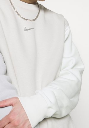 Osoba ubrana w biały sweter Nike z srebrnym naszyjnikiem w kształcie łańcuszka, z założonymi ramionami na tle jednolitego białego tła.
