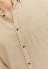 Beige Hemd mit Knopfverschluss aus strukturiertem Stoff, ausgestattet mit einer Tasche, braunen Knöpfen und einem dezenten Streifenmuster.