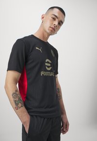 Black football shirt with textured pattern, gold accents, and red side panel. Features logo and branding. Model wears dark shorts.