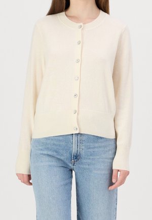 Femme portant un cardigan crème boutonné à manches longues et un jean taille haute bleu clair sur un fond uni.