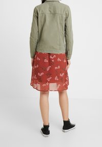 Chaqueta de denim verde sobre un vestido rojo con flores, de textura transparente y estampado botánico, combinado con zapatillas negras altas.