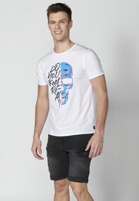 White short-sleeve t-shirt featuring a colorful graphic of a skull with text, paired with dark cargo shorts. Plain background.