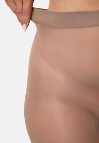 Persona che tira su la vita di collant trasparenti color carne sull'anca contro uno sfondo bianco semplice.