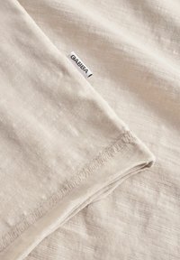 Beža auduma ar smalku tekstūru un mazu GABBA etiķeti; austs pārklājums izceļ gaismas variācijas krāsā un rakstā.