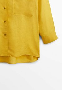 Chemise jaune à manches longues avec boutons, poche poitrine et manches avec poignets, montrée de la taille jusqu'à la fin des manches.