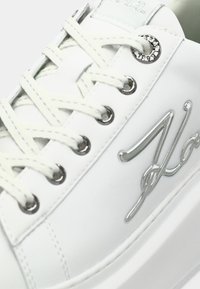 Close-up de um ténis branco com atacadores brancos, apresentando ilhós de metal prateado e um logótipo prateado em script de lado.
