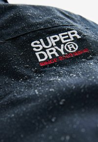 Giacca impermeabile navy con logo "SUPER DRY" in rilievo ricamato in bianco e rosso, caratterizzata da una superficie testurizzata con gocce d'acqua.