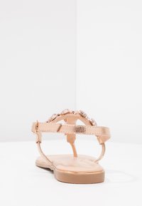 Rose gold sandal with a textured metallic strap and clear gem embellishments, flat sole, and adjustable ankle strap.