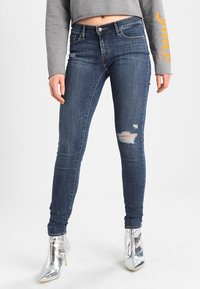 Woman wearing dark ripped skinny jeans, cropped gray sweatshirt, and shiny silver high-heeled boots, standing against white background.