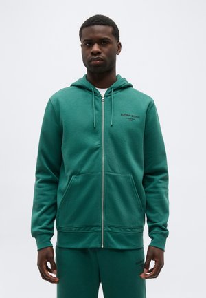 Schwarzer Mann trägt eine grüne Hoodie-Jacke mit Reißverschluss und passende Jogginghose mit Björn Borg Logos auf Brust und Oberschenkel, steht vor weißem Hintergrund.