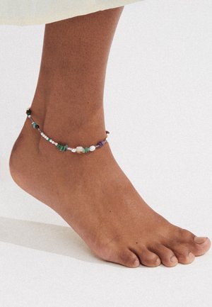 Enkel met een veelkleurige kralen enkelband met kleine steentjes en parels op een gladde huid tegen een lichte achtergrond.