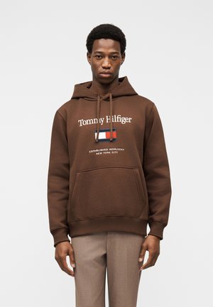 Młody mężczyzna w brązowej bluzie z kapturem Tommy Hilfiger z logo oraz beżowych spodniach, stojący na tle jednolitego białego tła.