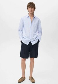 Camicia a maniche lunghe azzurro chiaro e bianca a righe, pantaloni corti blu navy e scarpe beige. La camicia ha un colletto con bottoni e una vestibilità comoda. Tessuto morbido.