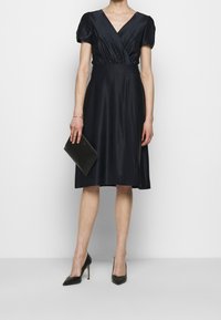 Žena v černých saténových šatech s krátkými balónovými rukávy, držící černou kabelku typu clutch, oblečená v černých špičatých lodičkách.