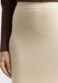 Beżowa dzianinowa spódnica ołówkowa o gładkiej fakturze i obcisłym kroju, z talią o średnim wzroście. Ręka delikatnie dotykająca tkaniny z boku.