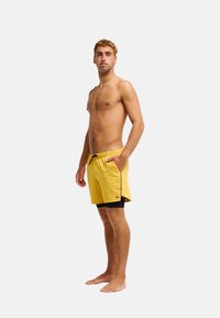 Homme torse nu portant un short de bain jaune avec une doublure intérieure noire, debout pieds nus, légèrement de profil, devant un fond blanc uni.