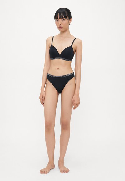 Vrouw die blootsvoets staat in een bijpassende zwarte bh en slip met merk-elastieken tegen een plain witte achtergrond.