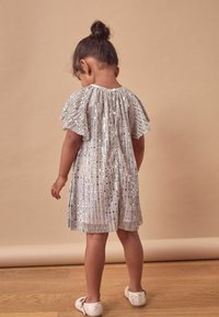 Silverfärgad veckad klänning med fladdrande ärmar och paljettdekoration. Barn som bär glittriga skor, står på trägolv mot en beige bakgrund.