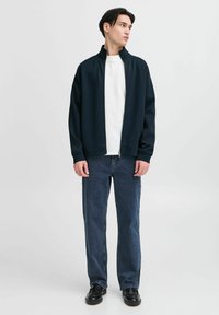 Marineblå zip-up jakke med høy krage, kombinert med en hvit T-skjorte og mørkeblå rette bukser, brukt med svarte sko. Glatt stofftekstur.
