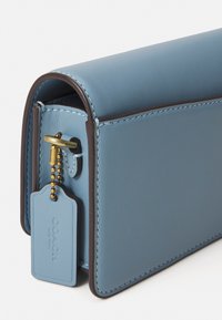 Borsa a tracolla in pelle azzurro chiaro con pattina arrotondata, bordi marroni e portachiavi in metallo dorato. L'etichetta attaccata presenta in modo prominente il logo "COACH".