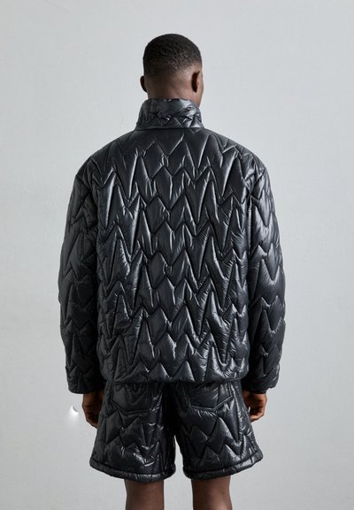 Schwarze steppierte Pufferjacke mit hohem Kragen und angularer Struktur, kombiniert mit passenden Shorts. Glattes, glänzendes Material.