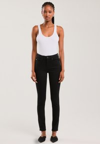 Naine seisab, kandes valget varrukateta topi, musti kõrge vöökohaga kitsaid teksaseid ja musti sussikujulisi kingi ühtlase tausta ees.