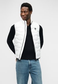 Νεαρός άνδρας που φοράει λευκό μοτίβο γιλέκο πάνω από μαύρο μπλουζάκι μακρυμάνικο και μπλε τζιν, στέκεται με τα χέρια πίσω από την πλάτη, χαμογελώντας ελαφρώς.