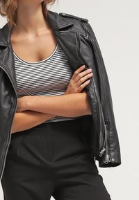 Schwarze Leder-Motorradjacke mit Reißverschlusstaschen, getragen über einem hellgrauen und weißen gestreiften Tanktop. Schwarze hoch taillierte Hose sichtbar.