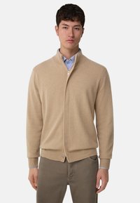 Beige zip-up cardigan i mjukt stickat material. Har hög krage, ribbad mudd och en knapppdetalj vid nederkanten.