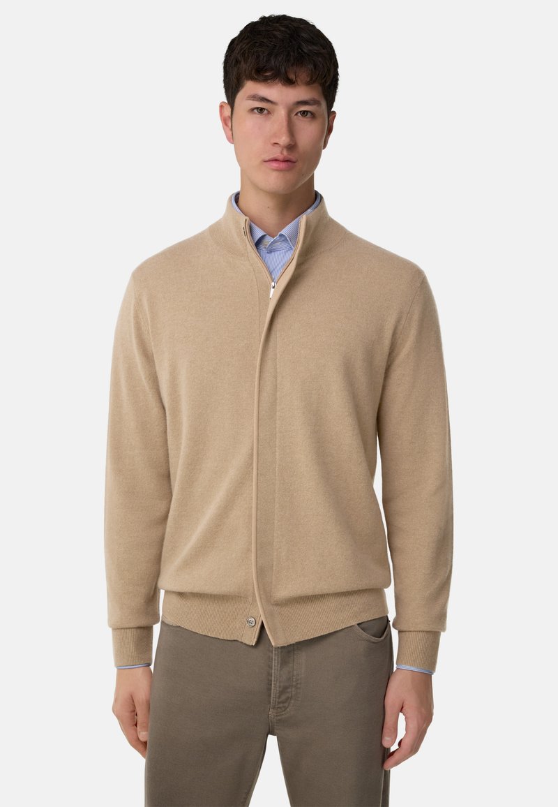 Beige zip-up cardigan i mjukt stickat material. Har hög krage, ribbad mudd och en knapppdetalj vid nederkanten.